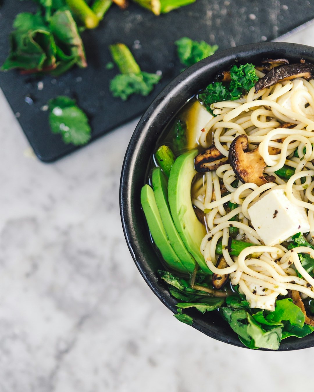 Vegan noodle soup with avocado and tofu in a black bowl.
